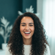 A smiling young woman, in her 20s, with a warm expression, against a soft, blurred background with deep teal and white tones.